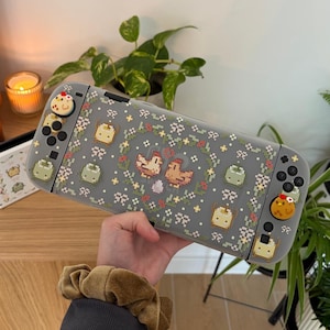 May include: A gray Nintendo Switch case with a pixelated floral and chicken design. The case has custom buttons with food-themed designs. A matching case is visible in the background.