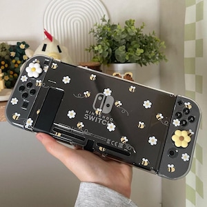 Può includere: Una console Nintendo Switch nera con una custodia trasparente che presenta un motivo di margherite e api bianche. La custodia ha il logo Nintendo Switch e le parole "Nintendo Switch" stampate su di essa. La console ha copri pulsanti gialli con un design a margherita.