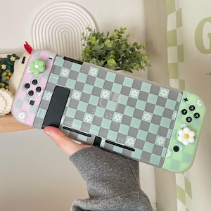 Può includere: Una custodia trasparente per Nintendo Switch di colore verde e rosa con un motivo a scacchi e piccoli fiori bianchi. La custodia ha un pulsante a forma di fiore verde sul lato sinistro e un pulsante a forma di fiore bianco sul lato destro.