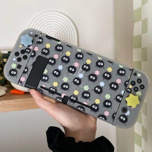 Puede incluir: Una funda protectora transparente para una consola Nintendo Switch con un estampado en blanco y negro de pequeñas criaturas redondas y esponjosas con ojos y un patrón de estrellas. La funda tiene un fondo gris claro y las criaturas están rodeadas de estrellas de colores pastel.