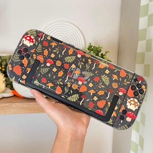 Könnte beinhalten: Eine schwarze und transparente Nintendo Switch-Hülle mit einem Muster aus Pilzen, Blättern und anderen herbstlichen Elementen. Das Design umfasst rote, orange und braune Farben. Die Hülle wird in einer Hand gehalten.