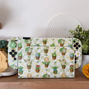 Könnte beinhalten: Eine weiße Nintendo Switch-Konsole mit einem individuellen Skin mit einem sich wiederholenden Muster aus Topfpflanzen und Zeichentrickfiguren auf einem gelb-grünen Karohintergrund. Die Controller haben grüne und weiße blütenförmige Knöpfe.