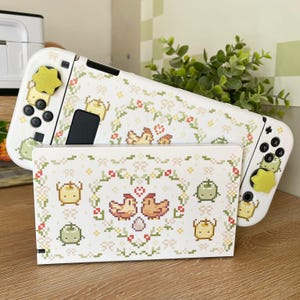 May include: White Nintendo Switch console with a custom design featuring a pixelated floral pattern and a couple of chickens. The console is in a stand with a matching design.
