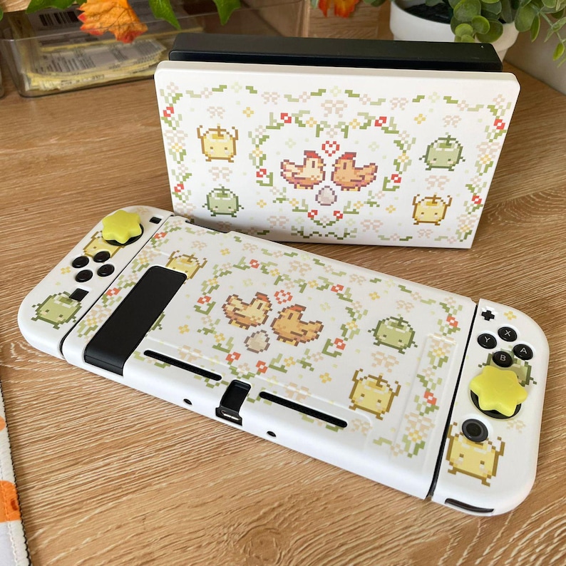 Puede incluir: Consola Nintendo Switch blanca con una piel personalizada que presenta un patr&oacute;n floral con pollos y huevos. La piel es blanca con acentos verdes, amarillos y marrones. La consola tiene una pantalla negra y botones amarillos.