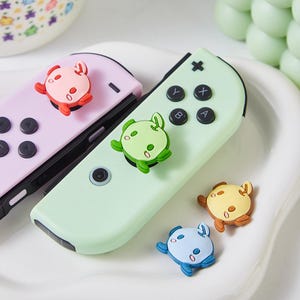 Könnte beinhalten: Vier bunte, cartoonartige Charakter-Knopf-Abdeckungen für einen Nintendo Switch-Controller. Die Charaktere sind ein rosa Krebs, ein grüner Frosch, ein gelber Krebs und ein blauer Krebs. Die Charaktere lächeln alle und haben große Augen.