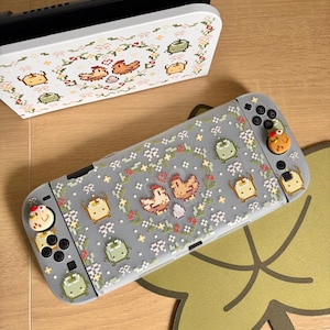 May include: A gray Nintendo Switch console and dock with a pixelated floral and chicken design. The console has a gray case with a pattern of chickens, flowers, and other pixelated shapes. The dock is white with a matching design.