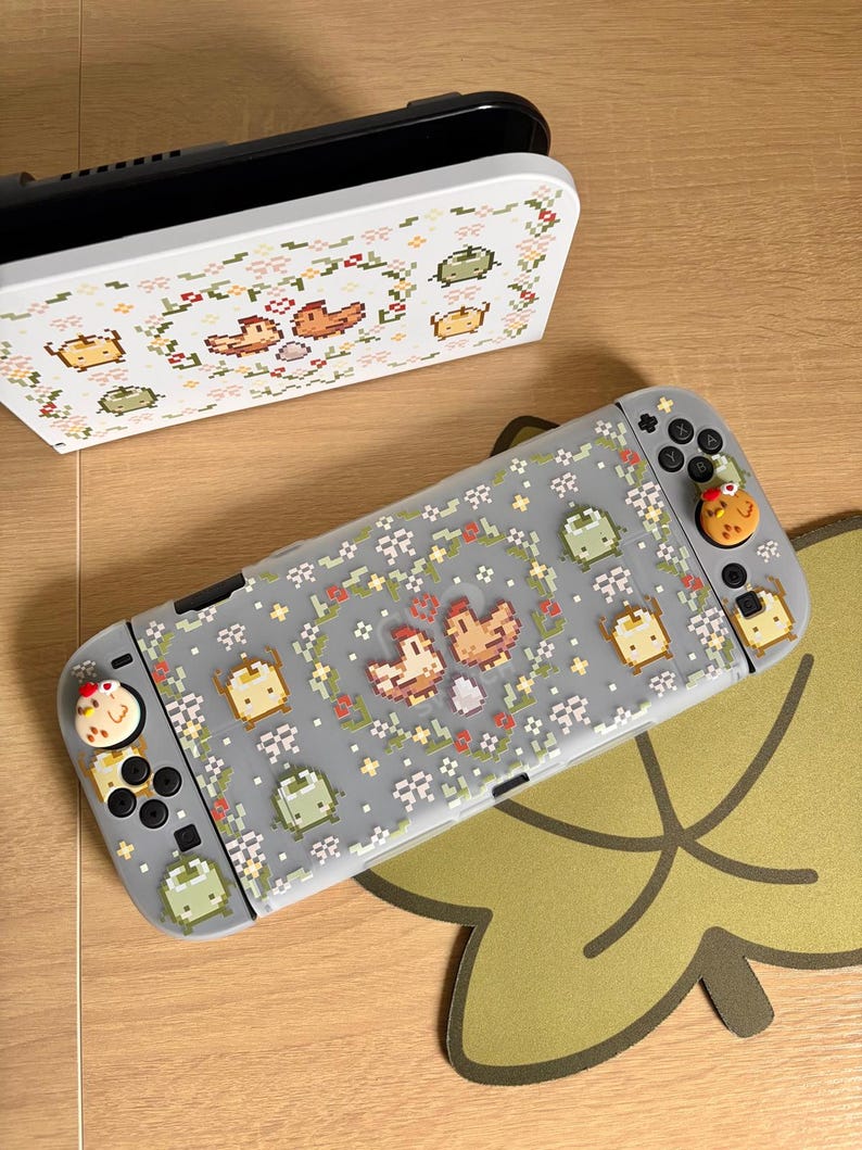 May include: A gray Nintendo Switch console and dock with a pixelated floral and chicken design. The console has a gray case with a pattern of chickens, flowers, and other pixelated shapes. The dock is white with a matching design.