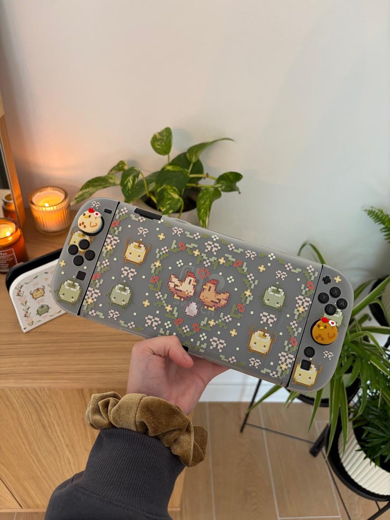 May include: A gray Nintendo Switch case with a pixelated floral and chicken design. The case has custom buttons with food-themed designs. A matching case is visible in the background.