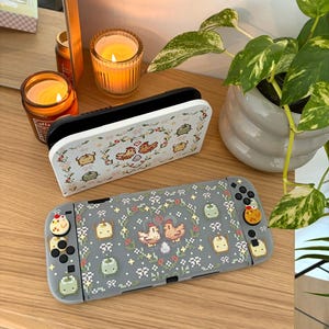 May include: A gray Nintendo Switch case and controller with a pixelated design featuring birds, flowers, and other small icons. The case and controller have a matching design. The controller buttons have decorative chicken-shaped toppers.