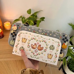 May include: A gray Nintendo Switch console with a matching case featuring a pixelated floral design and chicken graphics. The console has custom controller buttons with a chicken design. A white case with a chicken and floral design is in front.