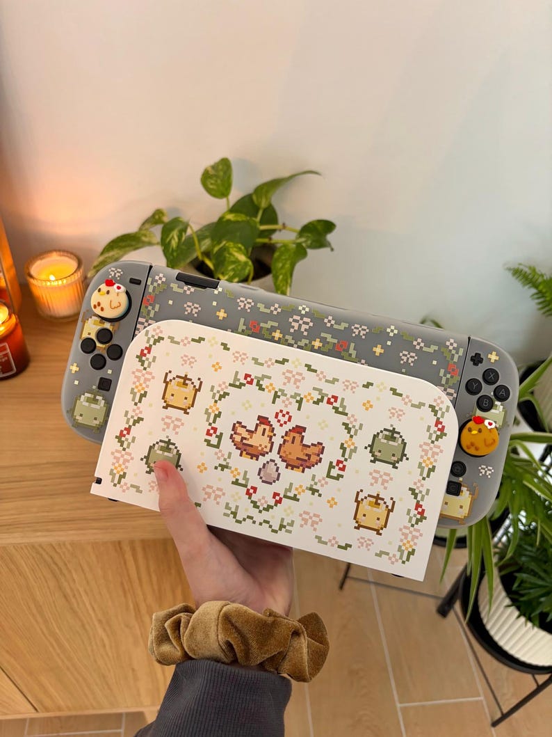 May include: A gray Nintendo Switch console with a matching case featuring a pixelated floral design and chicken graphics. The console has custom controller buttons with a chicken design. A white case with a chicken and floral design is in front.