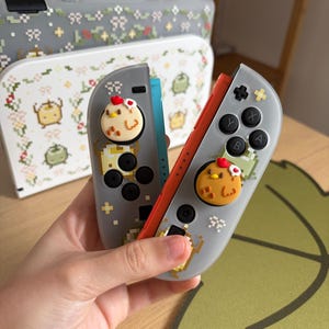 May include: Gray and red game controllers with pixelated designs and chicken-shaped button covers. The controllers have black buttons and a pixelated design. A matching case with a floral design is in the background.