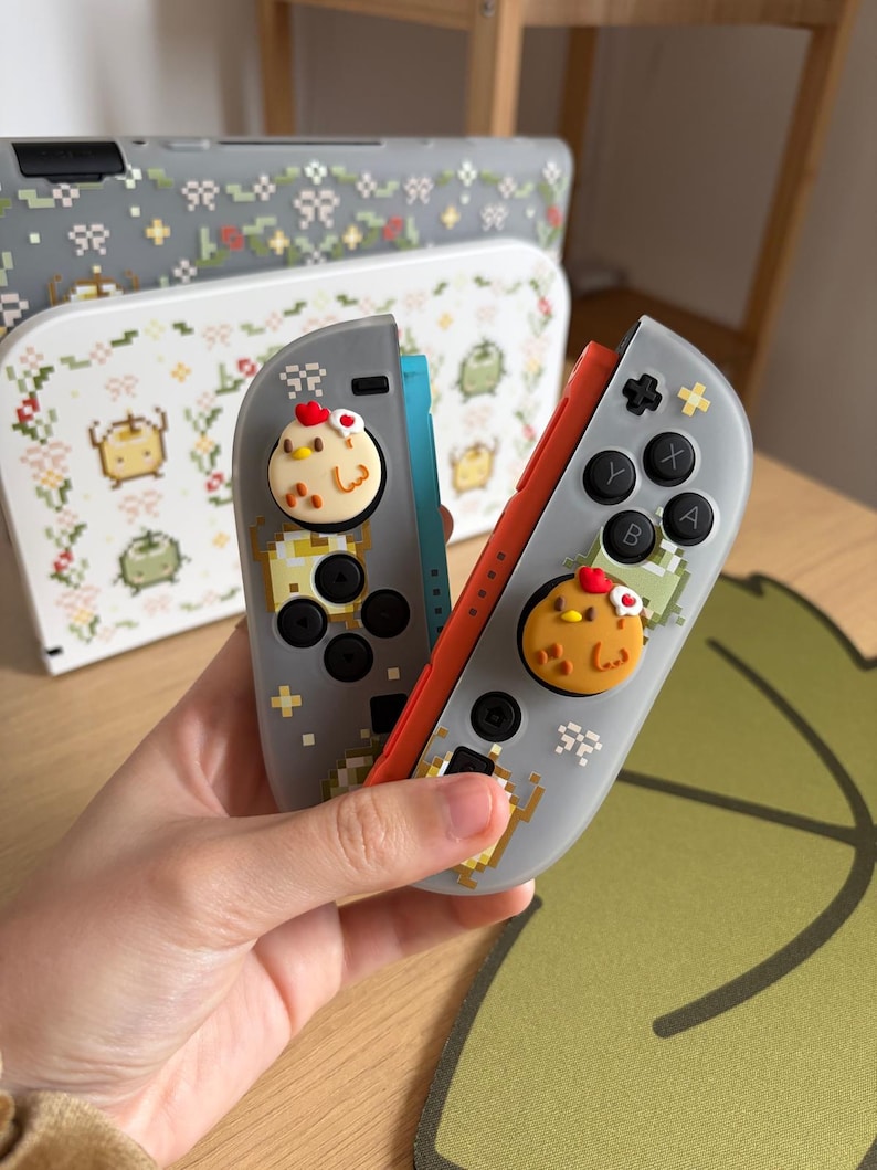 May include: Gray and red game controllers with pixelated designs and chicken-shaped button covers. The controllers have black buttons and a pixelated design. A matching case with a floral design is in the background.