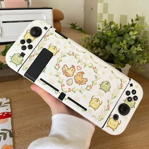 Puede incluir: Consola de juegos Nintendo Switch blanca con una piel personalizada que presenta un dise&ntilde;o pixelado de pollos y flores. La piel es blanca con acentos verdes y amarillos. La consola tiene un mando blanco y negro.