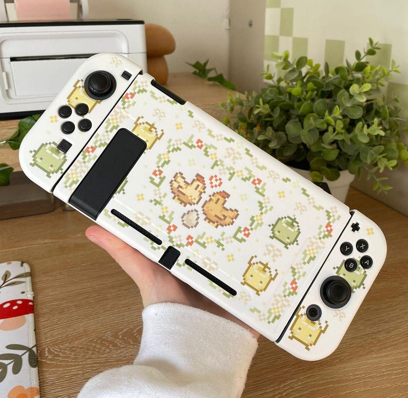 Puede incluir: Consola de juegos Nintendo Switch blanca con una piel personalizada que presenta un dise&ntilde;o pixelado de pollos y flores. La piel es blanca con acentos verdes y amarillos. La consola tiene un mando blanco y negro.