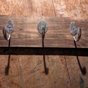 May include: A rustic wooden coat rack with three decorative metal hooks. The hooks are crafted from repurposed silverware, adding a unique touch. The wood has a dark stain, and the hooks are spaced evenly across the board.