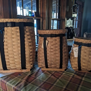 May include: Three woven baskets of varying sizes. The baskets are made of light brown woven material with black straps and trim. Each basket has a tag attached. The baskets are displayed on a patterned tablecloth.
