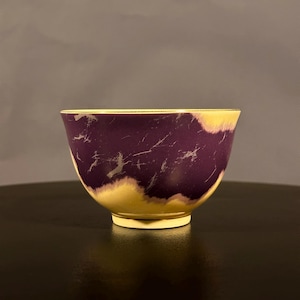 May include: A small, purple and cream ceramic bowl with a textured surface. The bowl has a gold rim and a slightly flared lip.
