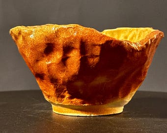 May include: A small, handmade ceramic bowl with a textured, uneven surface. The bowl is a warm, earthy brown color with hints of yellow.