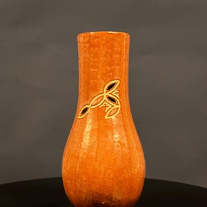 May include: Orange ceramic vase with a textured surface and a decorative gold and black leaf design. The vase has a bulbous shape and a narrow neck. The vase is a decorative home decor item.