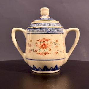 May include: A small white porcelain sugar bowl with a lid. The bowl has a blue and gold trim and a floral design in red, orange, and green.
