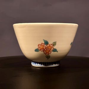 May include: A white ceramic bowl with a flared shape, featuring a hand-painted floral design in orange, red, and green. The base has a blue and white striped pattern. The bowl is likely used for serving food.
