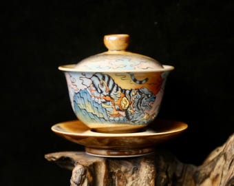 May include: A ceramic teacup and saucer set featuring a colourful tiger design. The cup has a lid and the saucer is brown. The tiger is depicted in a stylised, almost mythical way, with flames and clouds surrounding it.
