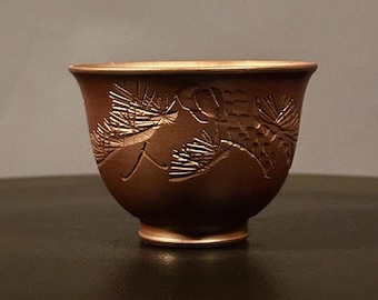 May include: A small, brown ceramic cup with a detailed, embossed pine branch design. The cup has a rounded shape with a slightly flared rim and a small, circular base. The cup is approximately 3 inches in diameter.