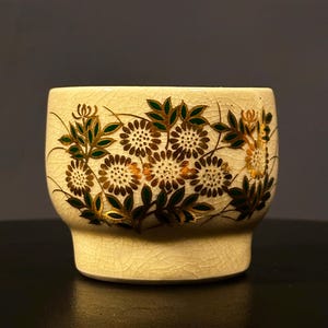 May include: Cream-colored ceramic cup with a floral design. The flowers are brown and gold, with green leaves. The cup has a crackled texture and a slightly flared base. The design wraps around the cup.