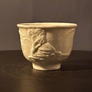 May include: A white ceramic cup with a raised design of a house and trees. The cup has a rounded shape and a small foot.