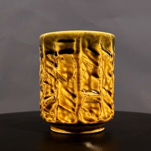 May include: A yellow ceramic mug with a textured, embossed design. The mug has a wide, rounded base and a slightly flared lip.