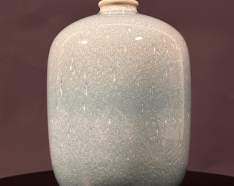 Może przedstawiać: Wazon ceramiczny z glazurą celadon i wykończeniem typu craquelé. Wazon ma zaokrąglony korpus i wąską szyjkę. Kolor jest blado-niebiesko-zielony, a powierzchnia ma sieć drobnych pęknięć. Wazon jest prawdopodobnie pochodzenia azjatyckiego.