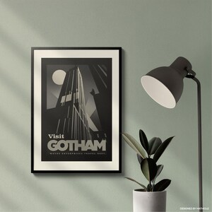 May include: A framed black and white art print with the text "Visit Gotham" and a stylized skyscraper design. The print is displayed on a light green wall, next to a black desk lamp and a potted plant.