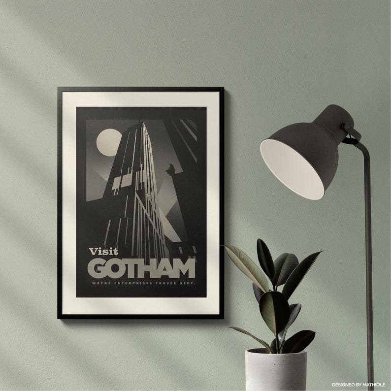 May include: A framed black and white art print with the text "Visit Gotham" and a stylized skyscraper design. The print is displayed on a light green wall, next to a black desk lamp and a potted plant.