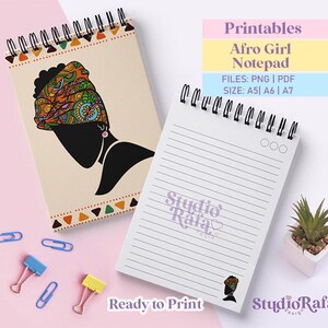 Peut inclure: Deux blocs-notes à spirales avec des pages lignées. L'un des blocs-notes présente une silhouette noire d'une femme avec un foulard coloré. L'autre bloc-notes porte le texte "Studio Rafa" et "Ready to Print". Des trombones colorés et une petite plante en pot sont également visibles.