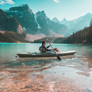 May include: A person kayaking on a turquoise lake with a mountain backdrop. The kayak is olive green, and the person is paddling with a black oar. The water is clear, revealing a rocky lakebed. Snow-capped mountains and a blue sky complete the scenic view.