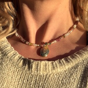 Puede incluir: Un collar de oro con un colgante redondo que presenta un dise&ntilde;o intrincado. El collar est&aacute; hecho de peque&ntilde;as cuentas de colores, incluyendo rosa, blanco y verde.