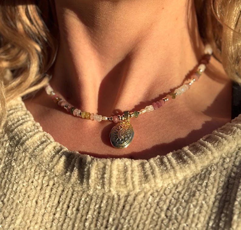 Puede incluir: Un collar de oro con un colgante redondo que presenta un dise&ntilde;o intrincado. El collar est&aacute; hecho de peque&ntilde;as cuentas de colores, incluyendo rosa, blanco y verde.