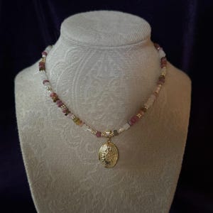 Puede incluir: Un collar de oro con un medall&oacute;n con un dise&ntilde;o floral. El collar est&aacute; hecho con cuentas rosas, blancas y marrones.