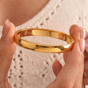 Könnte beinhalten: Ein polierter Armreif aus goldfarbenem Metall mit einer glatten, abgerundeten Außenseite. Die Innenseite des Armreifs ist mit dem eingravierten Text "Bitch Please, I'm Happy" in einer serifenlosen Schrift versehen.