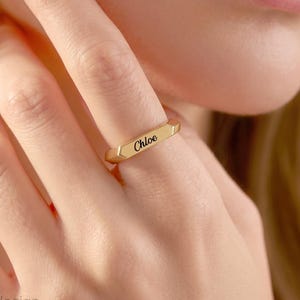Pode incluir: Um anel de metal dourado com uma superfície plana retangular. O nome Chloe está gravado em uma fonte cursiva preta no centro da barra. O anel possui um acabamento liso e polido com um design de banda fina e minimalista.