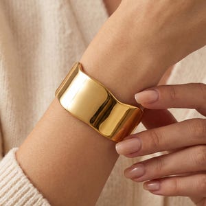 May include: A wide, polished gold cuff bracelet with a smooth, reflective surface and a slightly curved, undulating edge. The bracelet features a solid, metallic construction that wraps around the wrist, showcasing a bright, reflective finish.