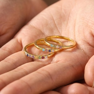 May include: Three thin gold rings, each featuring a row of small, round, multi-colored gemstones. The stones include shades of blue, pink, yellow, green, and purple, set into the polished gold bands. The text AGgiftsdesign is visible at the bottom.