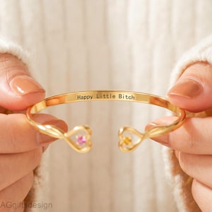 May include: A gold-toned open cuff bracelet featuring heart-shaped terminals, each set with a small gemstone—one pink and one yellow. The inner surface of the band is engraved with the black text "Happy Little Bitch" in a clean, sans-serif font.