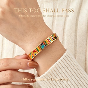 Könnte beinhalten: Ein goldfarbenes Armband mit einem lebendigen, mehrfarbigen Emaille-Muster aus geometrischen Formen in Rot, Gelb, Grün und Blau. Der Text "THIS TOO SHALL PASS" und "Internally engraved hidden inspirational sentence" ist darüber zu sehen.