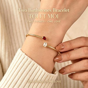 Könnte beinhalten: Ein goldfarbenes offenes Armband mit zwei verschiedenen Edelsteinen: einem birnenförmigen tiefroten Stein und einem rechteckigen klaren Stein. Der Text "Two Birthstones Bracelet TOI ET MOI TWO STONES ONE LOVE" ist im Hintergrund zu sehen.