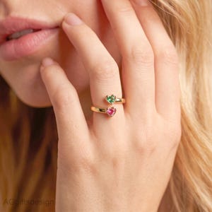Puede incluir: Un anillo de oro tipo bypass con dos piedras preciosas en forma de corazón, una verde y otra rosa, situadas en cada extremo de la banda abierta. El anillo se lleva en un dedo, mostrando el acabado de metal dorado pulido y los colores vibrantes.