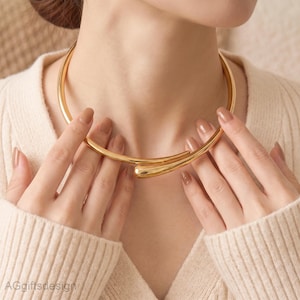 Pode incluir: Um colar tipo gargantilha de metal dourado com acabamento liso e polido e design aberto. O colar consiste numa banda rígida e circular que se estreita em duas extremidades arredondadas em forma de gota que se sobrepõem ligeiramente à frente.