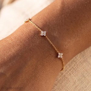 Könnte beinhalten: Ein zartes Goldkettenarmband mit gleichmäßig verteilten blumenförmigen Anhängern. Jeder Anhänger besteht aus vier kleinen, klaren, rund geschliffenen Steinen in einem Blumenmuster. Das Armband hat ein feines, poliertes Goldkettendesign.