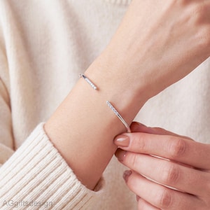 Può includere: Un bracciale aperto color argento caratterizzato da una sottile fascia con una fila di piccole pietre chiare e scintillanti incastonate su ciascuna delle due estremità. Il bracciale ha un design essenziale ed è indossato al polso.